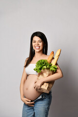 Young pregnant woman holding grocery paper shopping bag full of fresh vegetables and white bread. Diet healthy eating concept