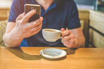 A man drinks coffee and writes on his smartphone while sitting at a table in a cafe