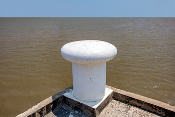 A shore with a white sea bollard on pier. The Large mooring bollard in a port with a blue horizon in background.