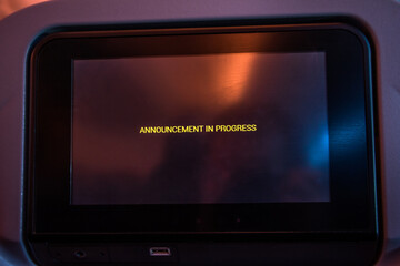 A monitor on backside of chair at airplane with text - announcement in progress - on display. Notice on screen in a flying aircraft.