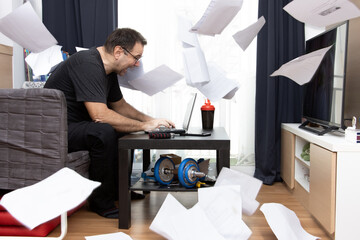 A dynamic zeal man sitting in living room and works on laptop, paper documents flying around. A...