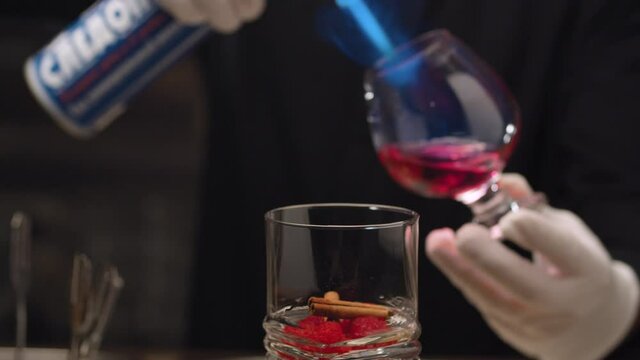 Making of Raspberry Sambuca. Close up barmans hand sets fire to sambuca in snifter and pours it into a glass with raspberries. Then he covers the glass. Steam rises from a drink. Glass slowly rotating