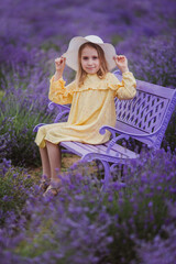 Smiling small girl in yellow dress and white hat sitting on violet bench in a lavender field