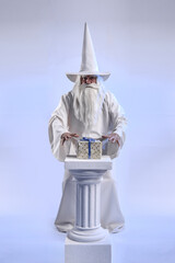 A grey-haired and stern wizard in a white cassock and a cap is doing sorcery and magic against a...