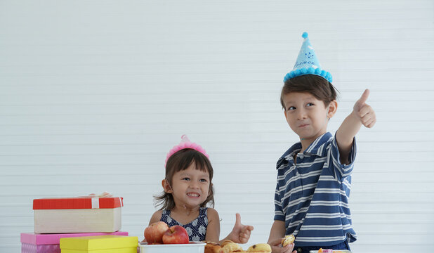 Birthday Party In Family. Little Caucasian Boy And Girl Thumbs Up Wear Cute Blue Pink Party Hat And Enjoy Eating Cookie, Jelly, Bread, Fruits, Apple In Birthday Party At Home 