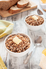Boiled buckwheat porridge in white round plates with slices of butter. in the background rye bread. On a light wooden background.