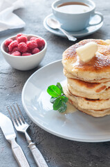 Pancakes with butter. A blue plate. Decorated with a sprig of mint. In the background is a cup with raspberries and a mug of tea. Close-up.