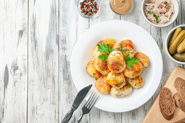 Roasted round potatoes with herbs and spices on a white plate. Next to the plate are pepper, mustard, sauerkraut, pickled gherkins and rye bread.