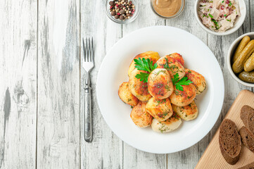Roasted round potatoes with herbs and spices on a white plate. Next to the plate are pepper, mustard, sauerkraut, pickled gherkins and rye bread.