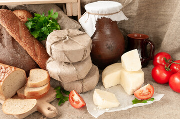 Adyghe cheese is cut into two parts. Nearby are three cheese heads packed in paper. Tomatoes, fresh bread, herbs.