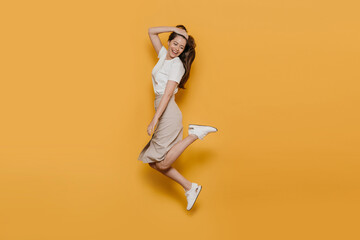 Fototapeta na wymiar Cheerful pretty brunette in white t-shirt, beige skirt and white sneakers, jumping high, careless mood, enjoying her life, ready for summertime. Studio shot over yellow backdrop. Happy people concept.