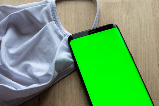Empty green display of a smartphone next to a white face mask on wooden surface. Ideal template for Corona Tracing App Mockup.