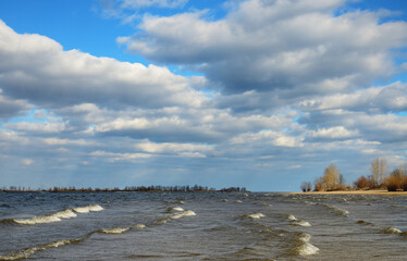 Landscape on the right bank of the Dnieper River in March, Cherkasy region, Ukraine. Clouds on a blue sky. Waves with white crests in shallow water.
