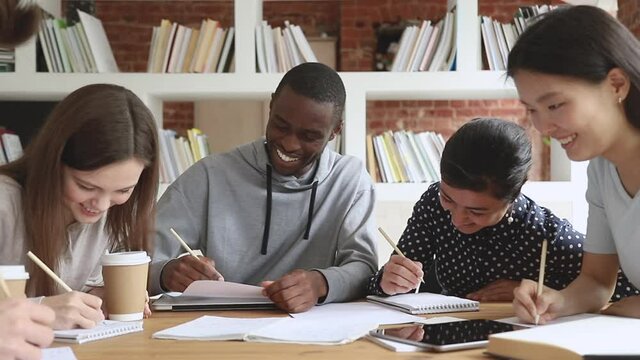 Cheerful multiethnic university college students team laugh at funny joke study together sit at table, happy diverse teen friends group having fun write homework work on project research in library