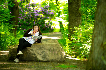 Lady sitting on a rock in a public square. She is reading a book and eating an apple.