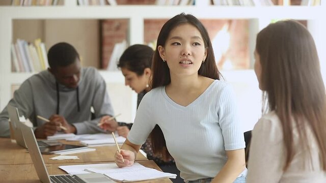 Diverse female college university students talking studying together in classroom, two asian and caucasian teen girls friends discussing high school assignment helping do research project in library