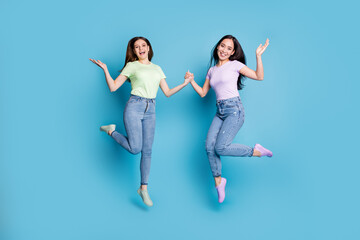 Fototapeta na wymiar Full length photo of two people lesbians couple ladies jump high up hold hands good mood rejoicing happy together wear casual t-shirts jeans footwear isolated blue color background