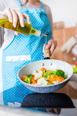 Close up of housewife holding bottle of olive oil and cooking pan with various vegetables in kitchen.