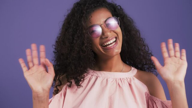 Afro-american woman in sunglasses and pink blouse. She smiling and waving her hands saying hi while posing against purple studio background. Close up