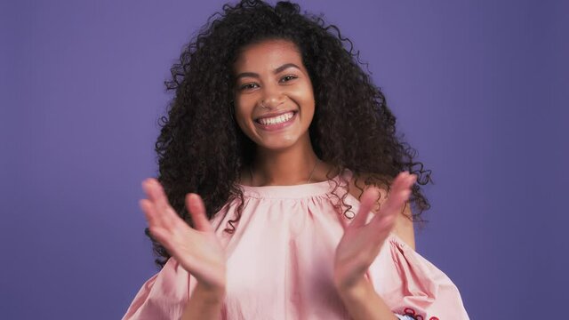 Dark-skinned woman in pink blouse. She throwing her head back demonstrating fluffy curly hair and smiling. Posing on purple background. Slow motion