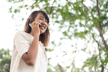 Happy Asian woman is using a mobile smartphone in a park on a blurry bokeh background of beautiful green trees