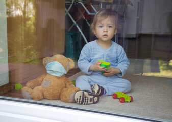 Child in home quarantine playing at the window with sick teddy bear wearing a medical mask against viruses during coronavirus COVID-2019 and flu outbreak. Photo made through the window