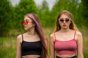 Two teenage girls with long loose hair and sunglasses pose as models against the backdrop of nature.