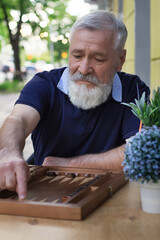 A man plays backgammon in a street cafe. Rest on a sunny summer day. Handsome elderly man with a beard