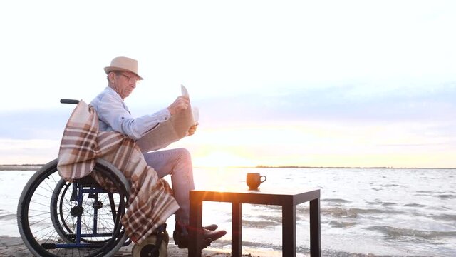 Disabled old senior grandfather reads newspaper sitting in wheelchair near water at sunset alone. Evening walk, comfortable shore relax, leisure of paralyzed adult pensioner handicapped man outdoors