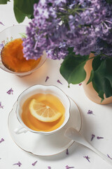 Bone china cup of tea with lemon, bowl with apricot jam and a vase with spring flowers (lilac) on white background