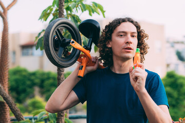 Young man on Father's Day with water pistol and a bicycle in hands, tired after playing with children, stands on the balcony in a blue T-shirt and jeans against the background of blurry palm trees