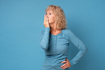 Scared middle aged senior woman covering mouth with hands feel horrified, stunned old blond female received shocking news. Studio shot on blue wall.