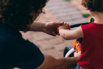 Father's Day. Dad with his little son on a bike from back. Family walk with a child. Outdoor activities for family. Dad helps son with a bike. Hands of father and small child.