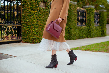 Detail of young fashionable woman wearing beige wool coat, tulle midi skirt and black high heel cowboy boots. She is holding stylish burgundy handbag in hands. Street style.