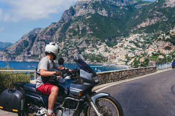 A motorbike traveler poses against the backdrop of an incredibly beautiful view of Positano, a city on the edge of cliffs, the sea and a winding road. Travel and tourism. Active lifestyle. Italy