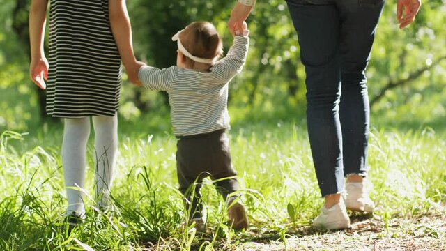Mother and elder sister walk with baby toddler making first steps on park grass happy family girls play together on nature