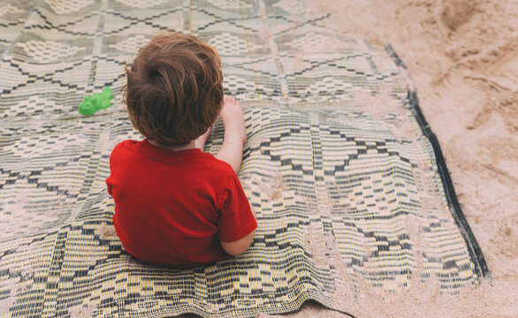 Father's Day. Little Son Sits Alone On The Beach And Waits For Dad. Happy Loving Family. A Lonely Child In Red T-shirt Sits On A Bedspread From The Back. Before You Go Swimming In The Sea