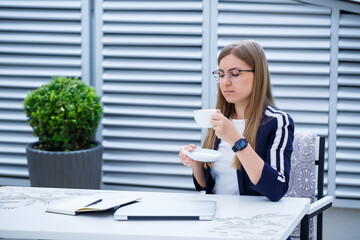 Beautiful young woman in a white T-shirt is drinking coffee and working on a laptop and smiling while sitting outdoors in a cafe. Young woman laptop for work. Female freelancer working on a laptop