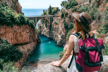 A girl in a cowboy hat with a backpack enjoys an incredibly beautiful view of a stone bridge over the gorge Fiordo di Furore. Little bay with paradise beach. Back view. Amalfi, Italy