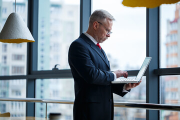 Smiling happy managing director thinks about his successful career development while standing with a laptop in his office near the background of a window with copy space