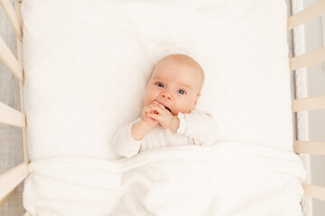 Portrait of a baby with his hands in his mouth in a white crib. Interior of the children's room and bedding for children. A cute little girl is lying in her crib. 