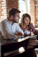 caucasian couple looking at laptop in front of them, illuminated by light of a monitor, talking, drinking coffee in cafe