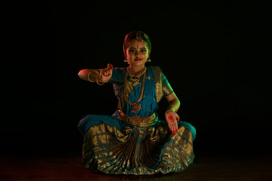Young bharatnatyam dancer sitting in muzhu mandi position while performing in front of a dark background. 
