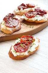 Toast (italian bruschetta) with sun-dried tomatoes and soft curd cheese with fresh herbs on wooden board, light background, closeup view, soft selective focus
