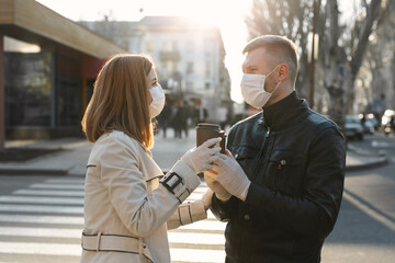 Young couple, a man and a woman in medical masks and gloves, drink coffee from disposable cups on the street and look at each other against. Coronavirus. Covid 19