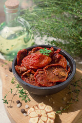 Homemade sun-dried tomatoes in oil with fresh herbs (thyme, oregano), garlic in a bowl on wooden board,  light background, soft selective focus