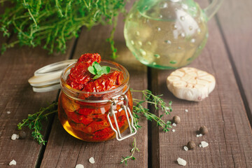 Homemade sun-dried tomatoes in oil with fresh herbs (thyme, oregano), garlic in a glass jar on dark wooden background , soft selective focus