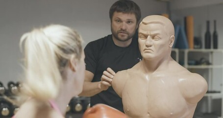 Back view of blond female boxer punching mannequin with blurred personal trainer talking at the background. Slim woman training in gym with equipment. Cinema 4k ProRes HQ.