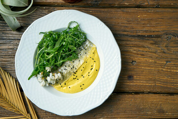 Appetizing fried seabass fillet with yellow sauce and arugula in a plate on a wooden background