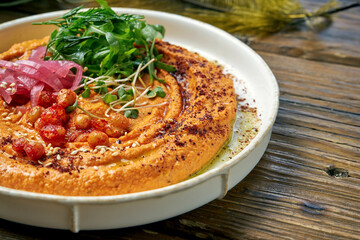 Classic oriental appetizer - baked peppers hummus with greens and arugula in a white plate on a wooden background